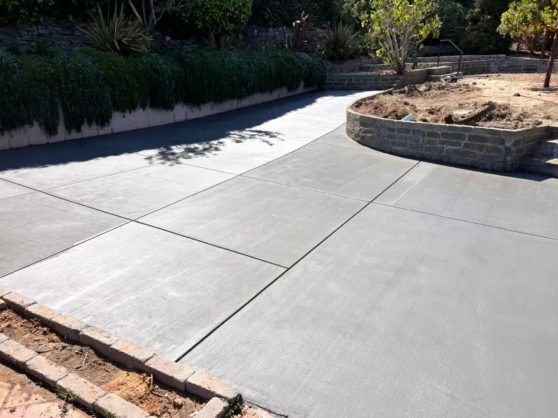 Gray concrete driveway with cut lines, leading towards a retaining wall and greenery.