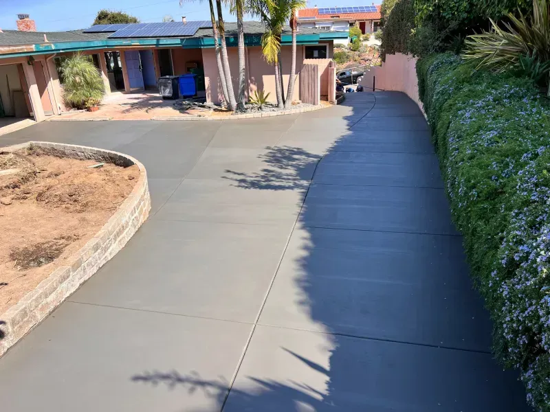 Newly poured, grey concrete driveway leading up to a home with a pink exterior, lined by green hedges.