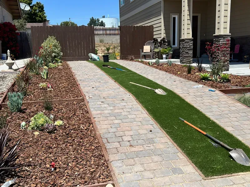 Driveway with brick pavers and artificial turf strip, lined with plants and mulch. Shovels rest on turf.