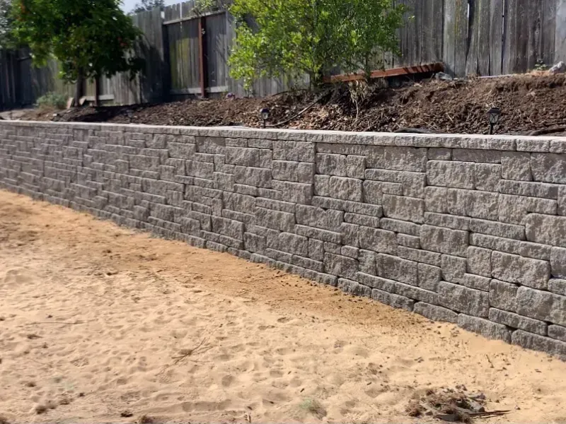 Gray retaining wall made of stacked blocks alongside a sandy area, with a wooden fence and trees in the background.