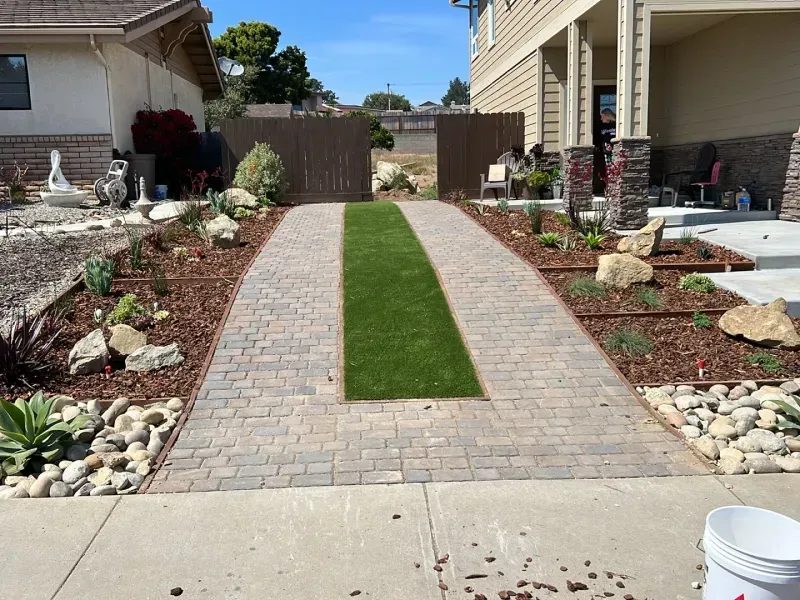 Driveway with brick pavers, artificial turf strip, and landscaped beds.