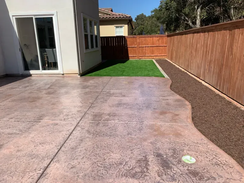 Backyard patio with stamped concrete, a patch of green turf, and a brown gravel border along a wooden fence.
