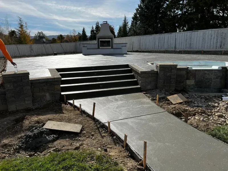 Freshly poured concrete patio with steps, pathway, and a fireplace. Construction site with fence in the background.