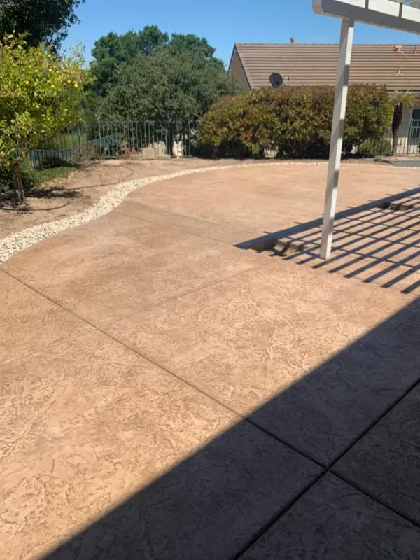 Brown stamped concrete patio with pergola shadow, curved rock border, and backyard greenery.