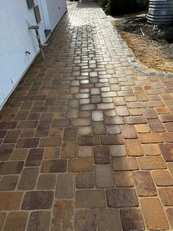 Brick pathway leading through a sunlit area, bordered by a white wall and a patch of dirt.