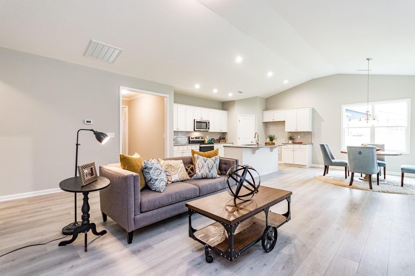 Open-concept living space: gray sofa, kitchen, dining table. Light wood floors, gray walls, white cabinets.
