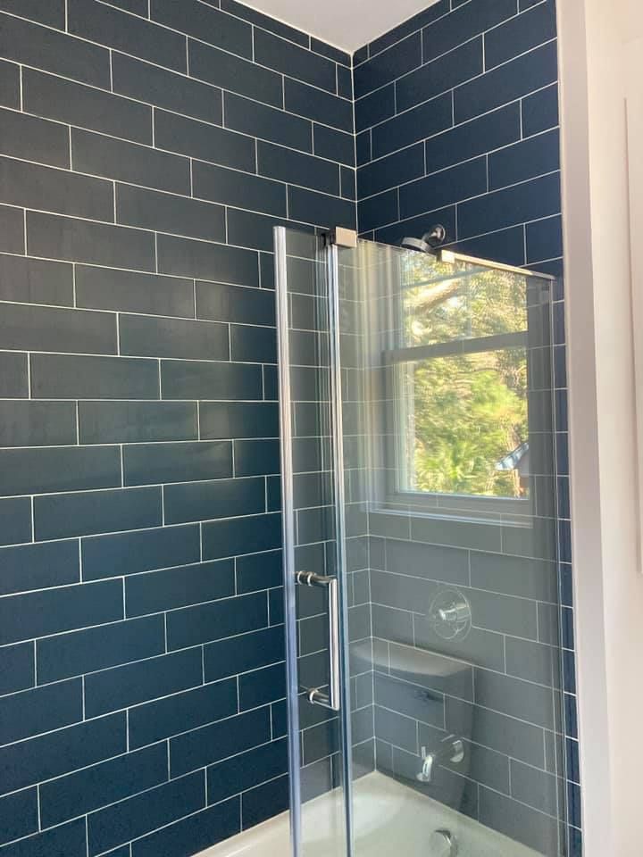 Dark blue tiled shower wall with a glass door. A window is reflected in the glass.