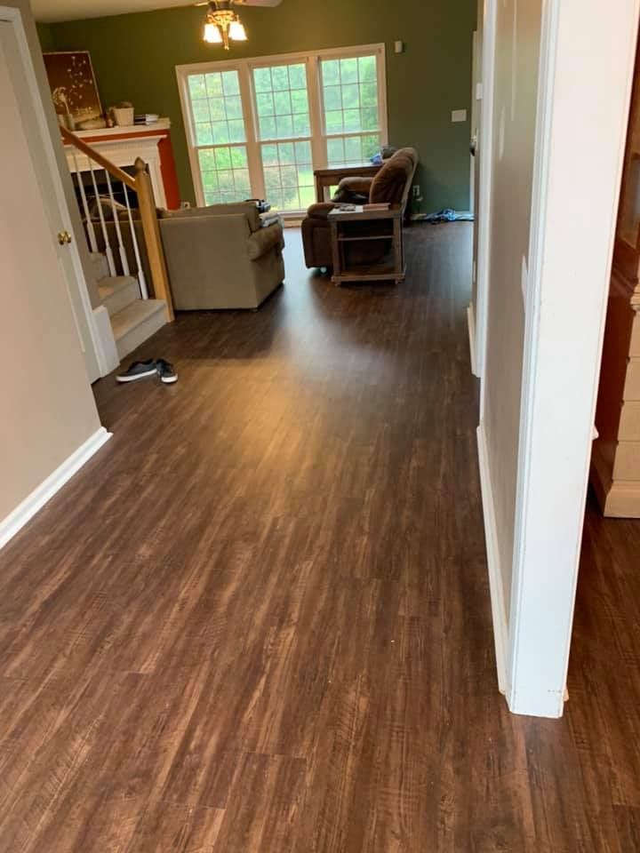 Dark wood-look flooring in a home, leading to a living room with a chair, and window.