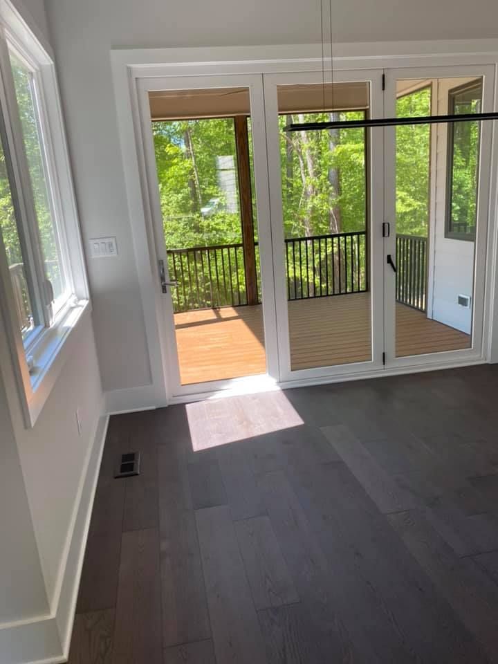 Interior room with dark hardwood floors, a sunny window, and glass doors leading to a wooden deck with lush greenery.