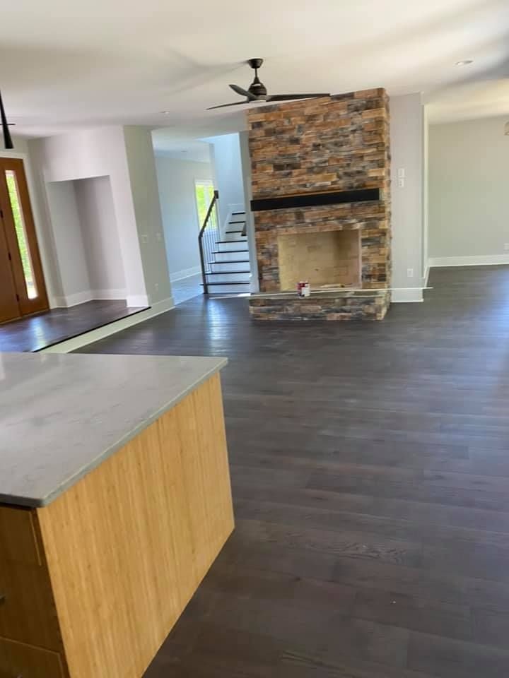 Open-plan living space with stone fireplace, dark wood floor, and light gray walls. A kitchen island is in the foreground.