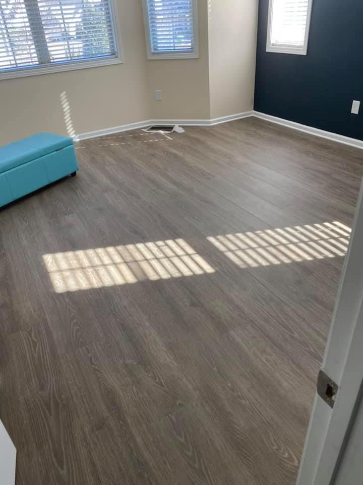Empty room with gray wood-look flooring, blue accent wall, and blue ottoman. Sunlight streams through windows.