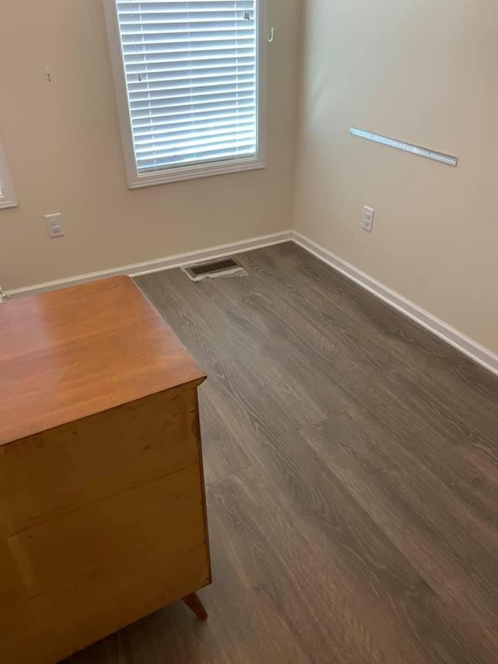 Bedroom corner with a wooden dresser, dark wood-look floor, window with blinds, and beige walls.