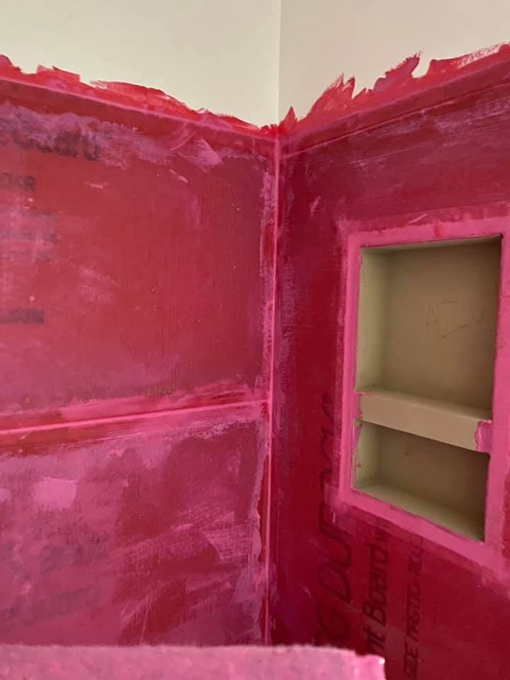 Red-coated shower walls with a recessed shelf, corner angle. Pink sealant around the edges, white ceiling.