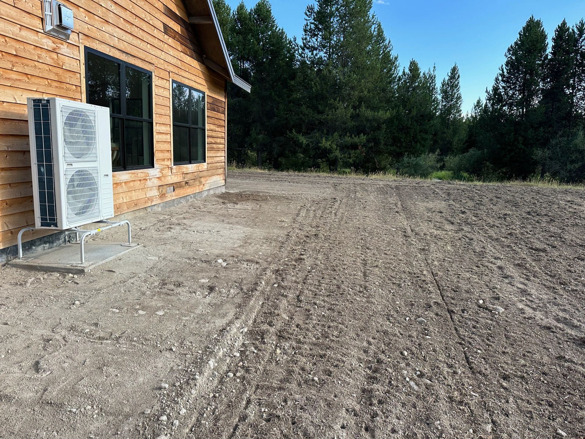 Graded yard beside modern home with natural wood siding, showing proper foundation clearance and utility installation with heat pump mounted on concrete pad