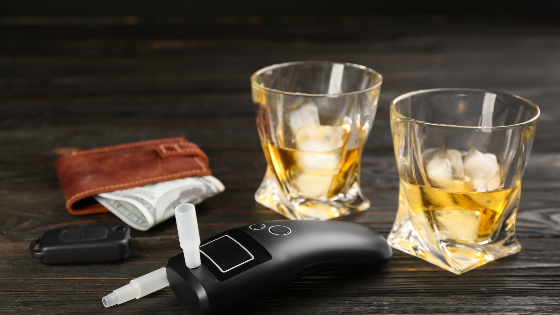 Two whiskey glasses with ice, a wallet, car keys, and a breathalyzer on a dark wood surface.