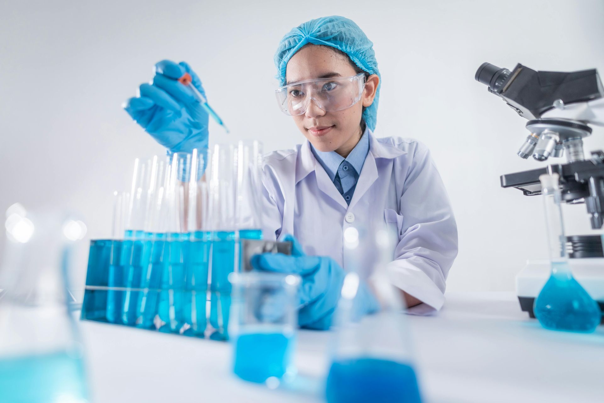 Scientist in lab coat and gloves using a pipette with blue liquid, working with test tubes, microscope in the background.