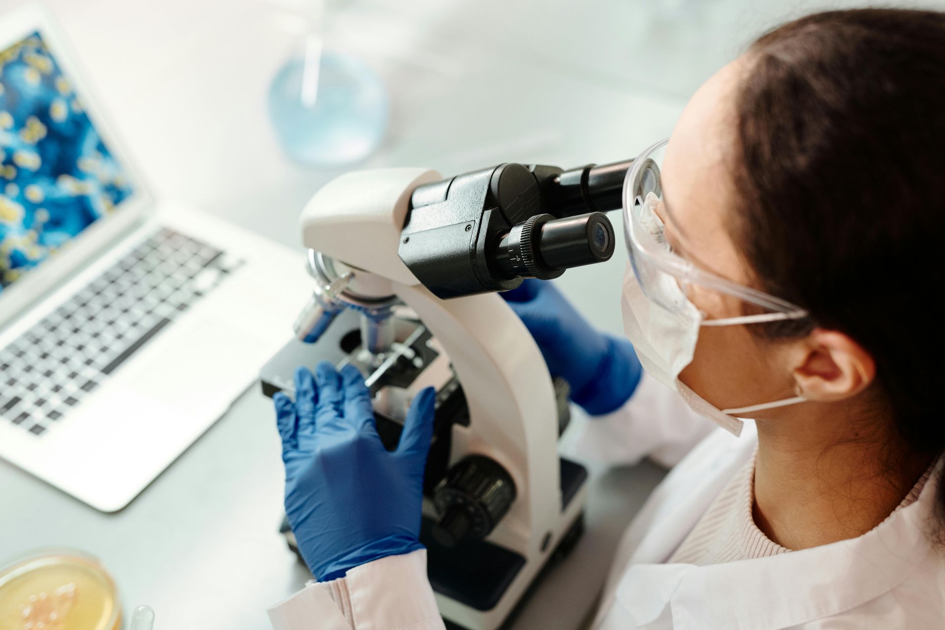 Person in lab coat, gloves, and mask using a microscope, laptop nearby, lab setting.
