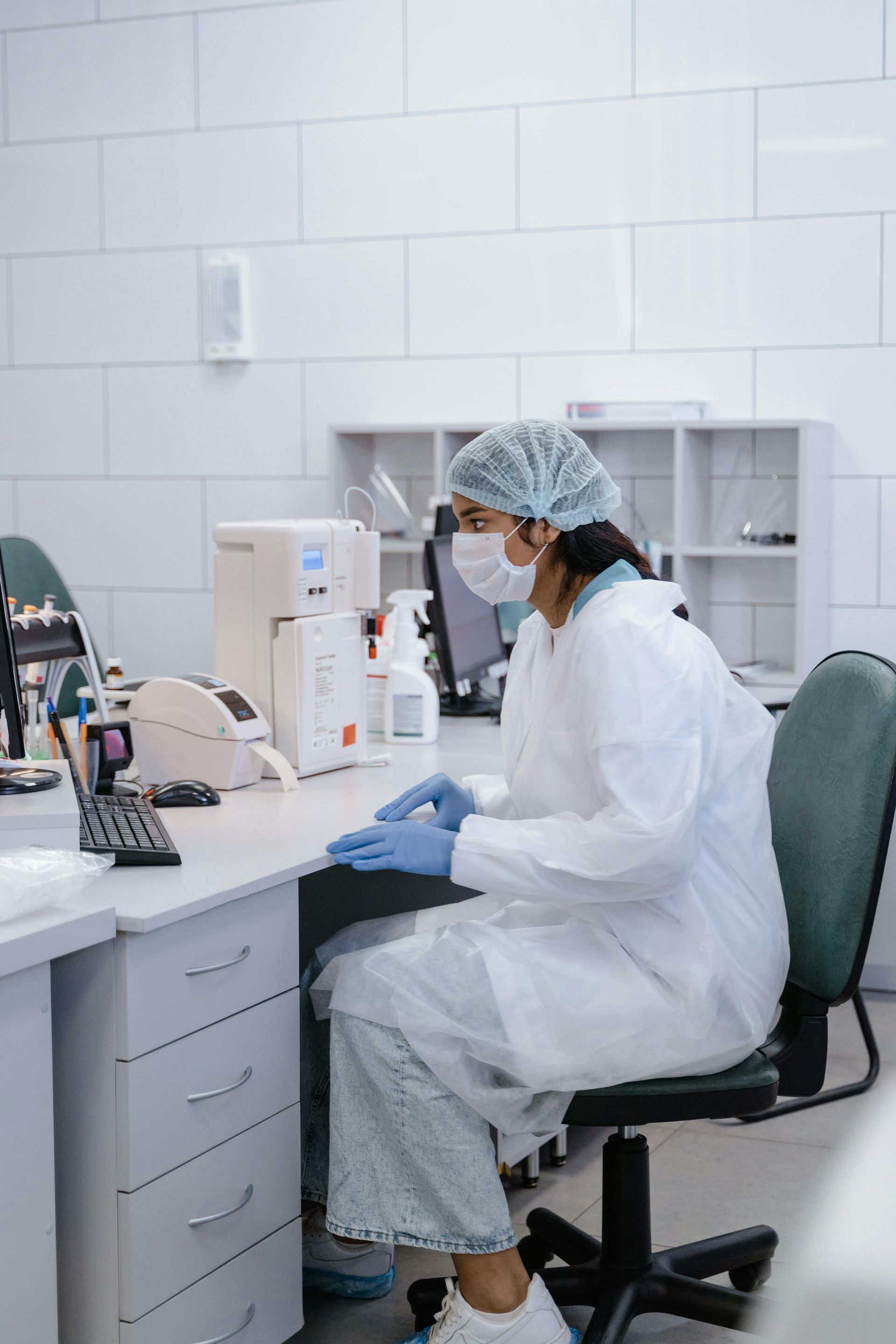 Lab worker wearing PPE sits at a desk, typing on a computer. Laboratory setting, white walls.