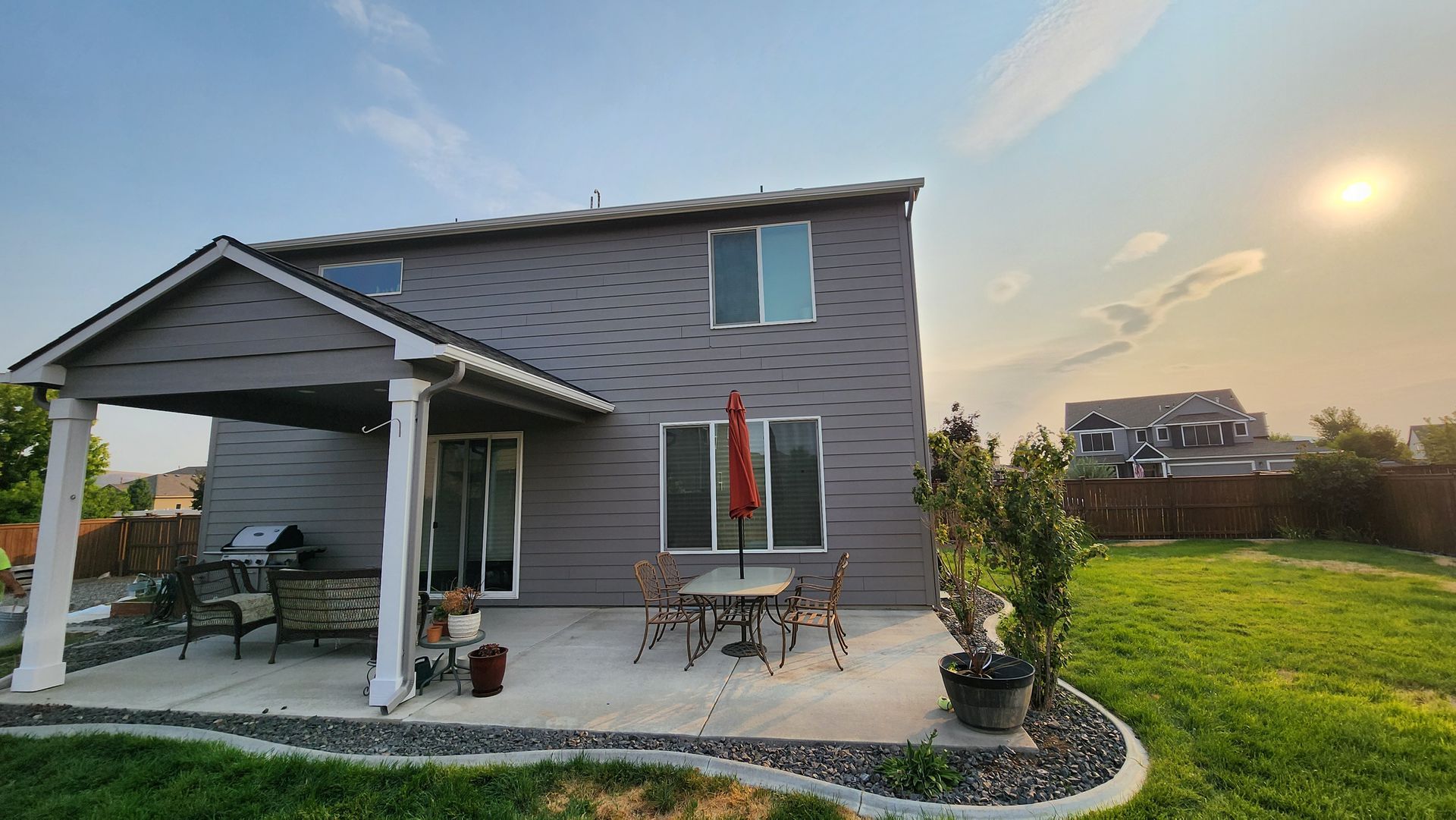 Two-story gray house with a patio, umbrella, and small table and chairs set, under a blue sky with the sun.