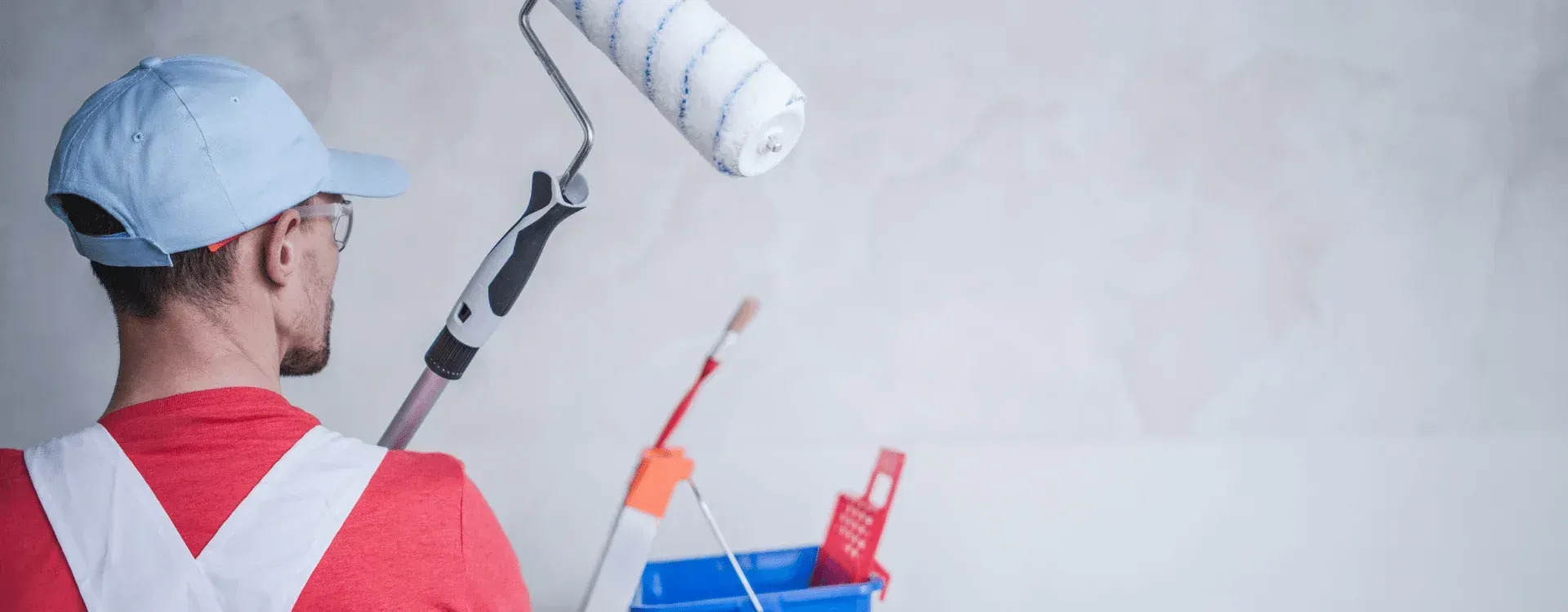 A person wearing a hat and red shirt with white suspenders uses a paint roller to paint a white wall.