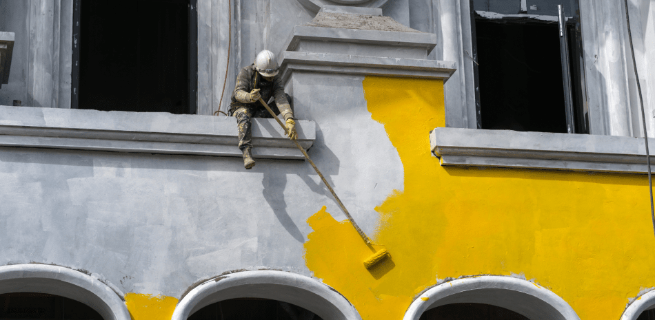 A street sculpture of a painter in a hard hat painting a building yellow. He is sitting on a ledge, with a roller extended towards the wall.