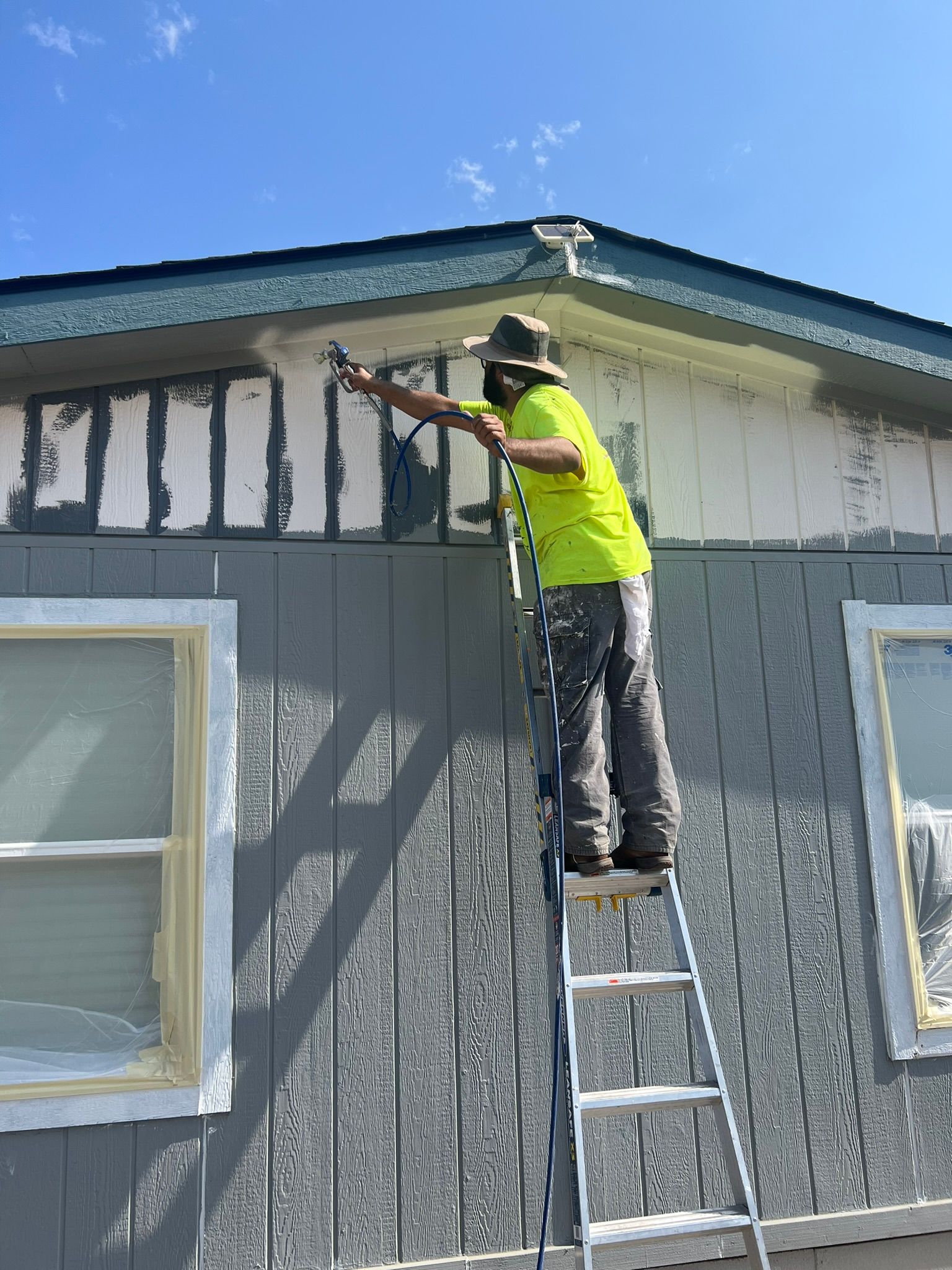 A person in a neon yellow shirt sprays paint on the side of a house, standing on a ladder.