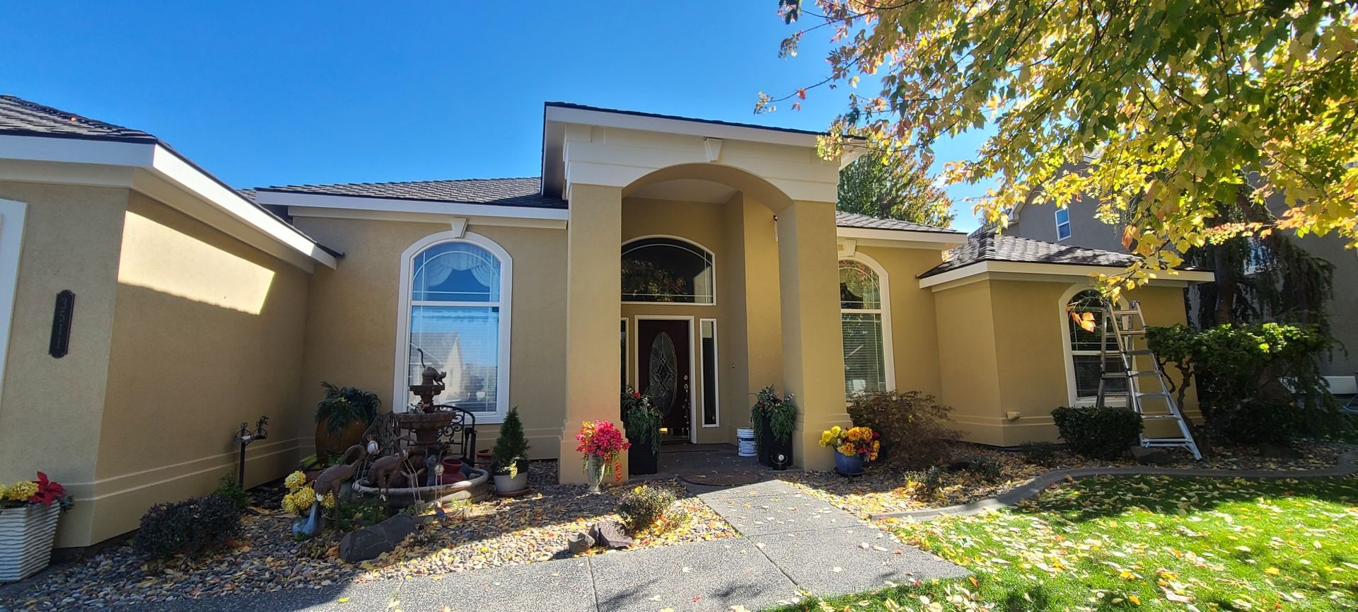 A house with beige walls, dark roof, and a stone walkway is surrounded by fall foliage under a blue sky.