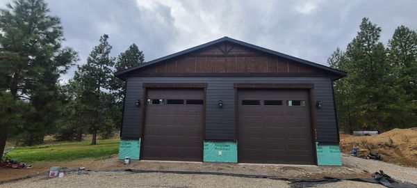 A two-car garage with brown doors and dark siding, set against a cloudy sky and surrounded by trees.