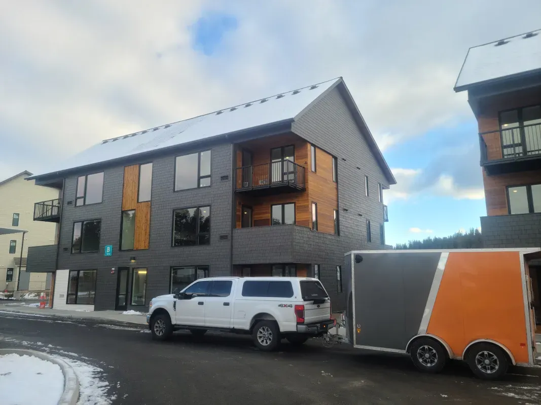 A white truck towing a gray and orange trailer parked in front of a modern building with gray and wood-tone siding, snowy ground.