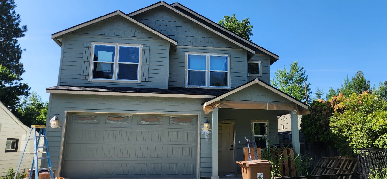 Two-story house with light green siding. A garage door and front porch are visible. Bright blue sky in the background.
