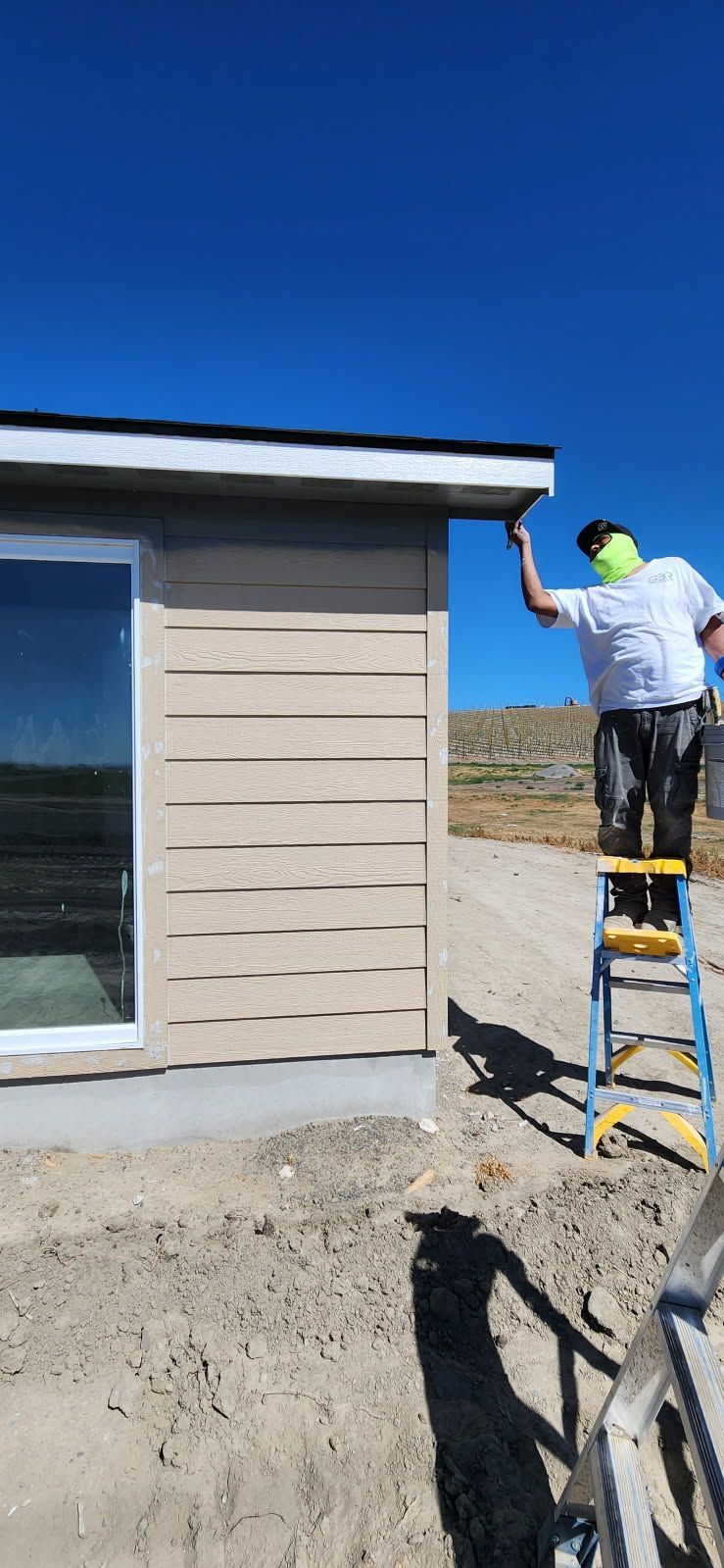 A person painting the eaves of a building while standing on a ladder on a sunny day. The sky is clear blue.