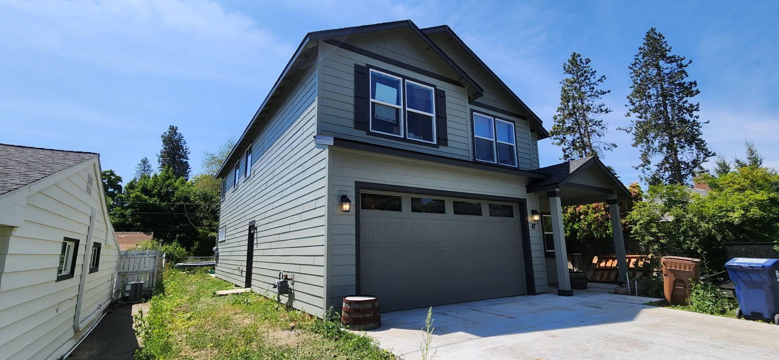 Two-story house with gray siding and a two-car garage on a sunny day. Adjacent building on the left, trees on the right, and a blue sky.