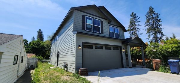 Two-story house with gray siding and a two-car garage on a sunny day. Adjacent building on the left, trees on the right, and a blue sky.