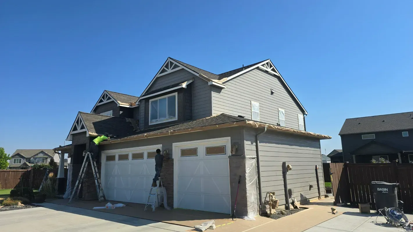 Two-story house with gray siding, white garage doors, and a worker on a ladder. Blue sky.
