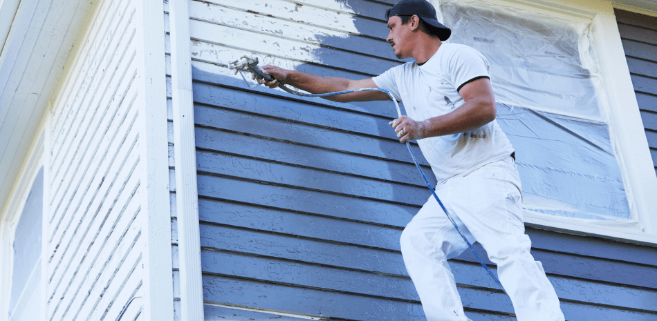 A person on a ladder sprays paint on the siding of a house, with the initial color blue and the new color white.