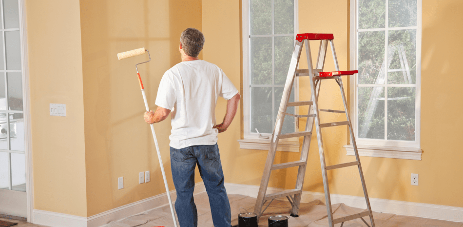 A person painting a wall inside a room. They are holding a paint roller with a ladder nearby.