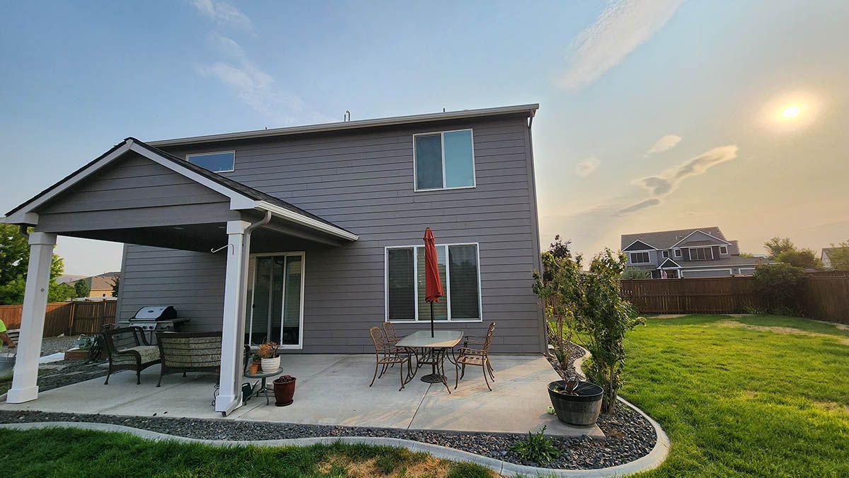 Backyard of a two-story gray house with a patio, table, chairs, and grill. Sunny day with a blue sky and a grassy lawn.