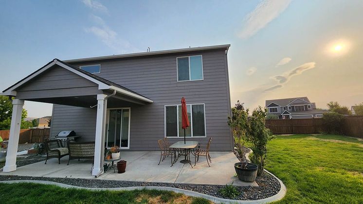 Backyard of a two-story gray house with a patio, table, chairs, and grill. Sunny day with a blue sky and a grassy lawn.