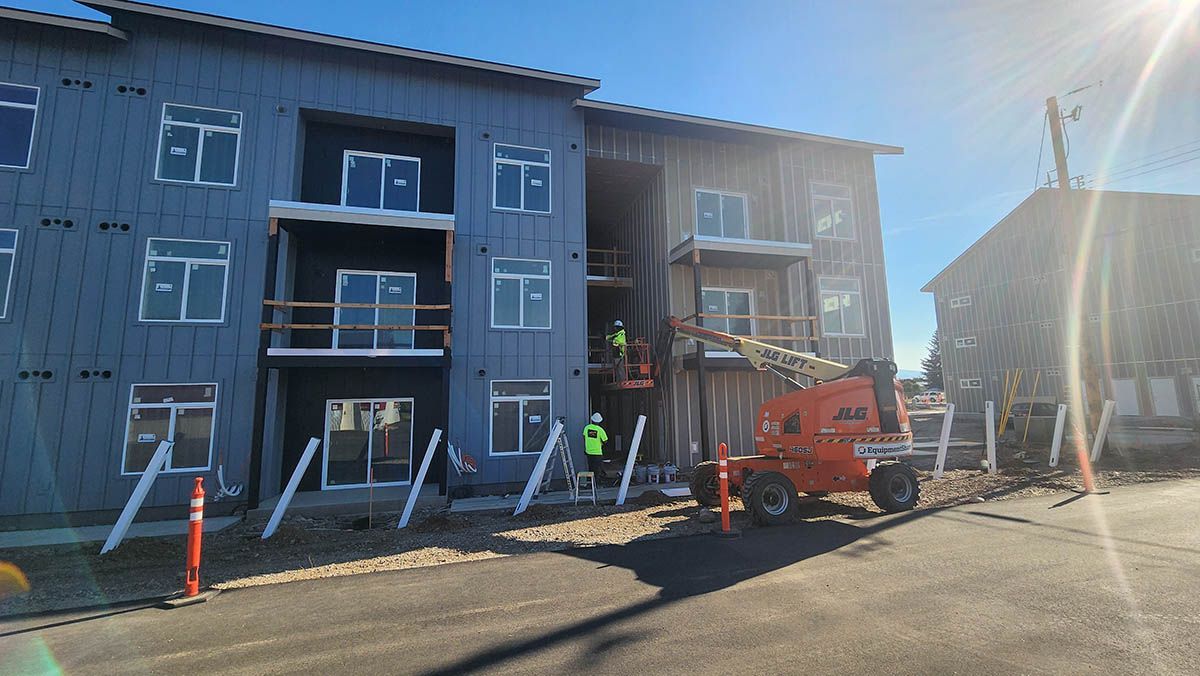 Apartment building under construction; workers on lift. Exterior walls have grey siding and balconies. Bright sunlight.