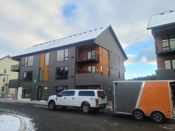 Apartment building under construction; workers on lift. Exterior walls have grey siding and balconies. Bright sunlight.