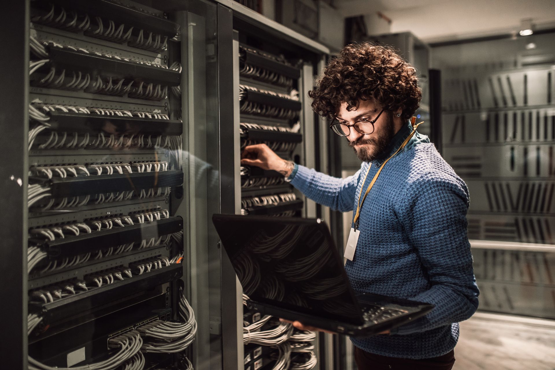Man with curly hair and glasses works on a server rack with laptop in a data center.