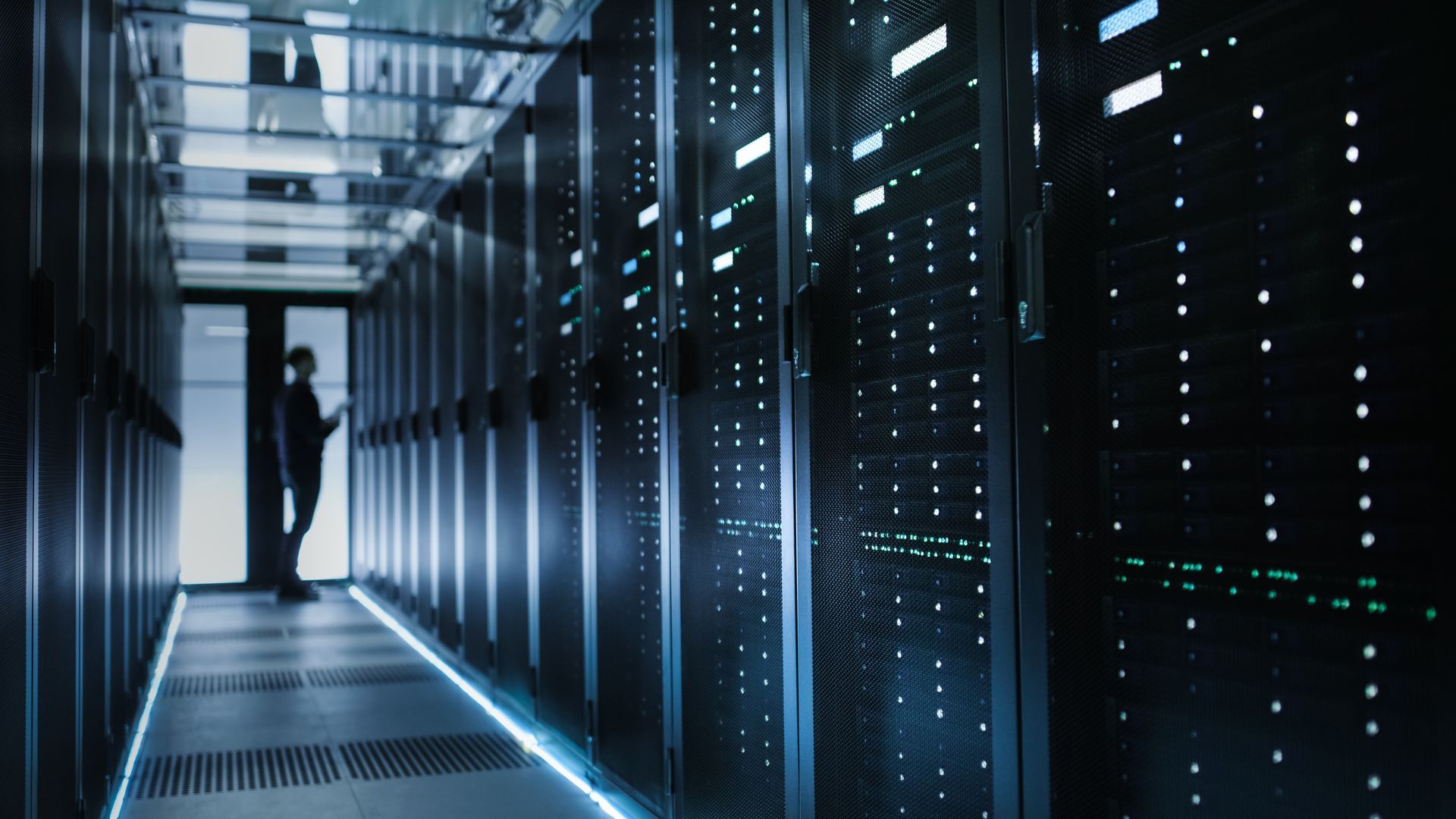 Person in a server room, standing near a row of data racks. Blue lighting illuminates the narrow aisle.