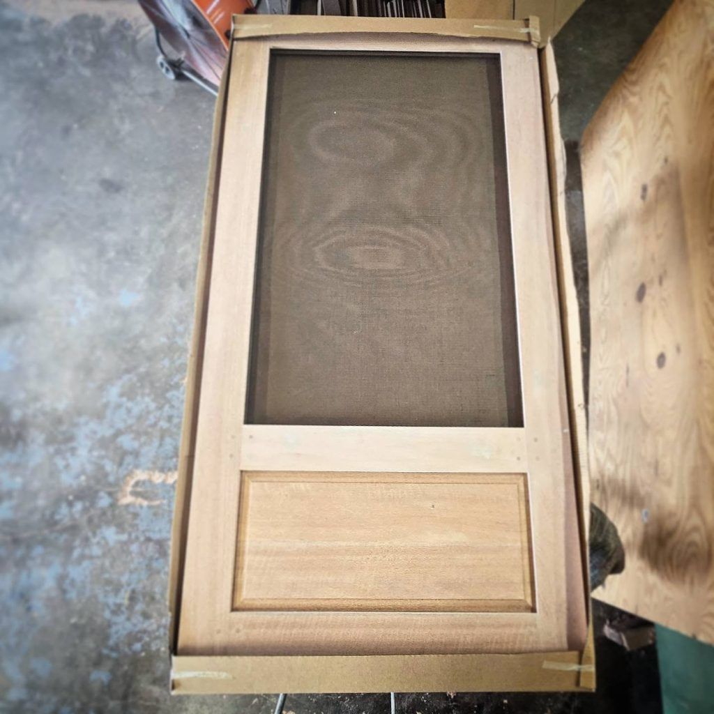 Unfinished wooden screen door in cardboard packaging, with a screen panel and lower solid panel.