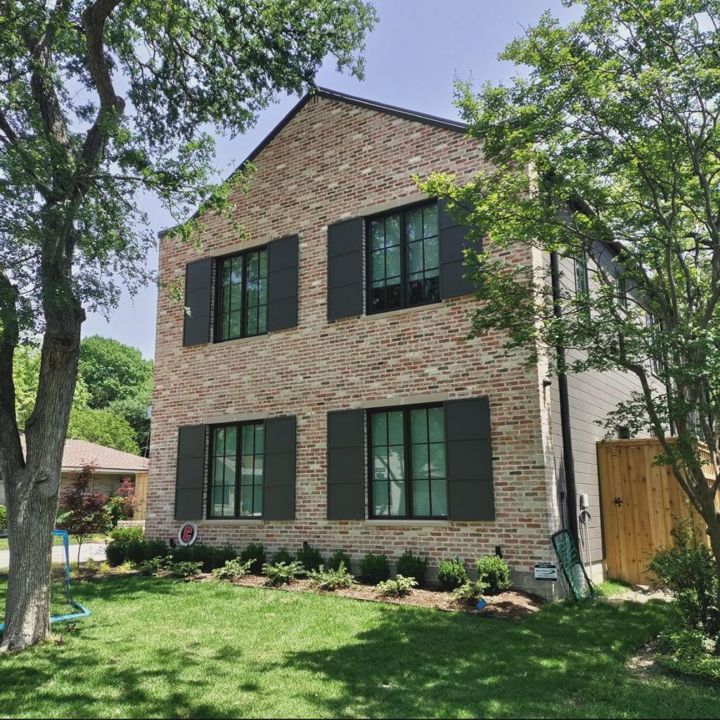 Two-story brick house with dark shutters, green lawn, trees, and blue sky.