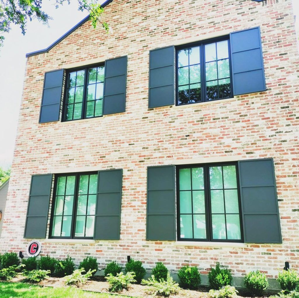 Two-story brick house with black-framed windows and dark gray shutters, shrubs in front.