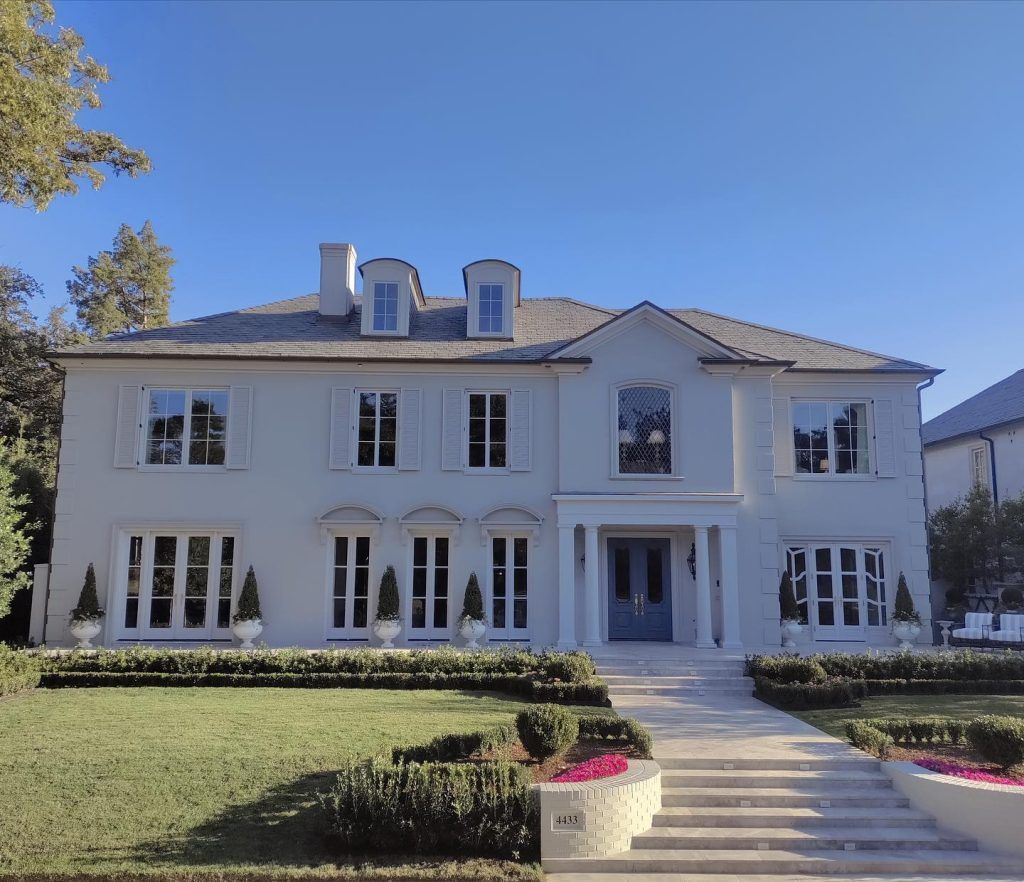 Two-story light blue house with multiple windows, blue door, and manicured landscaping under a blue sky.