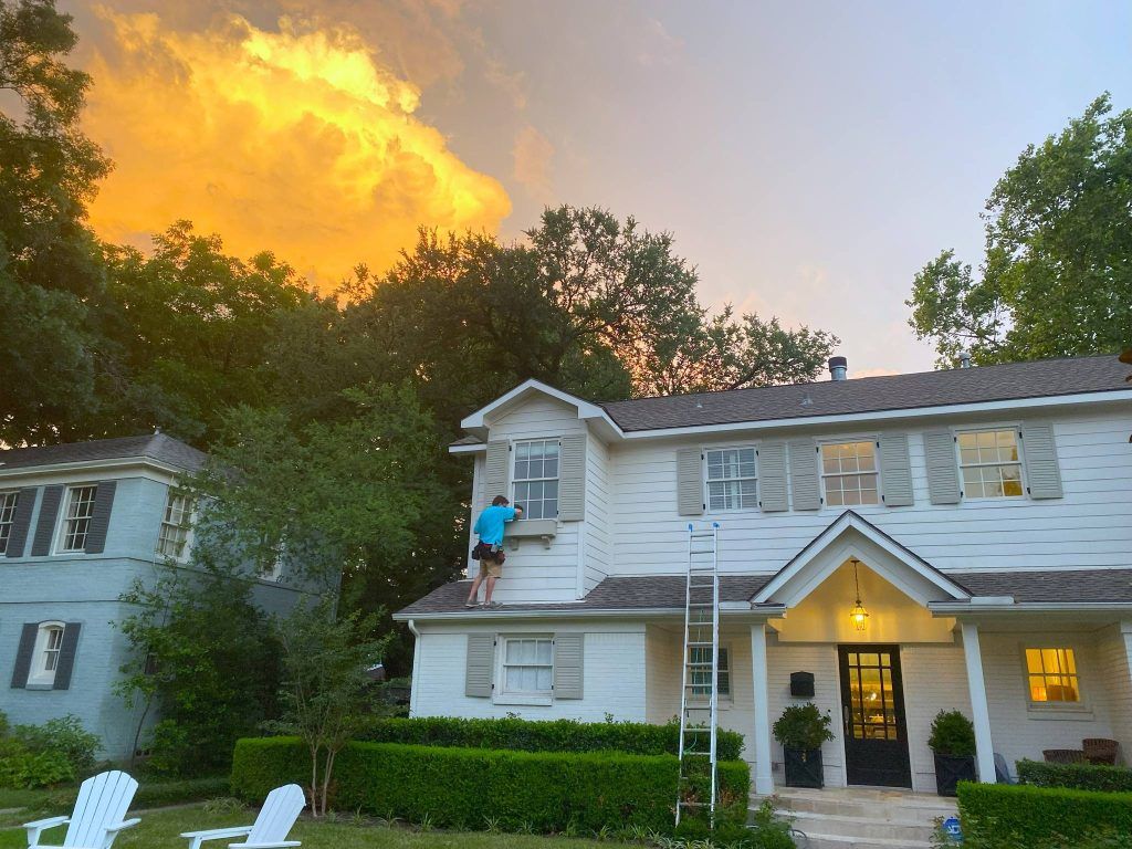 Person on ladder washing windows of a white house with a dramatic sunset in the background.