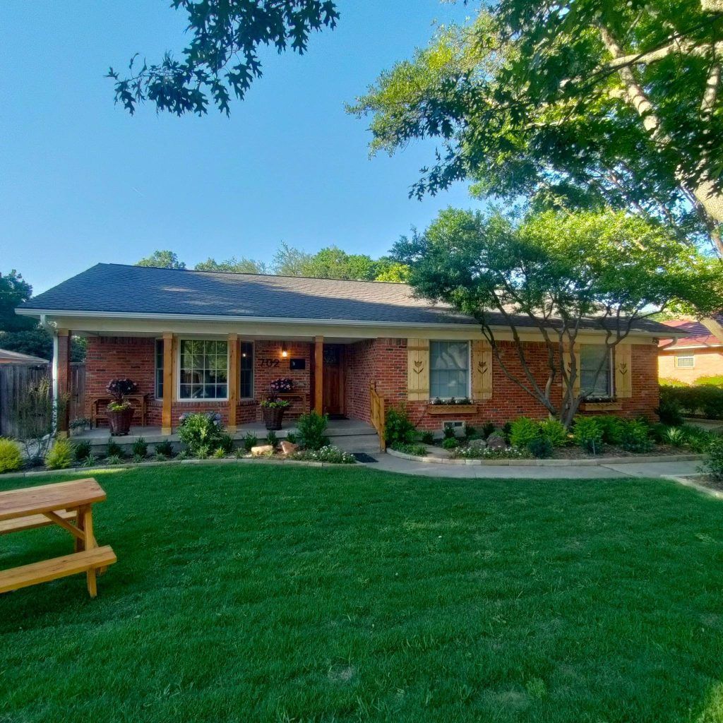 Brick house with a green lawn, porch, and trees. Yellow shutters and picnic table.