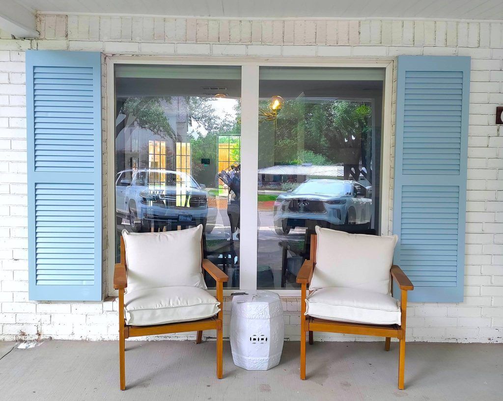 Two chairs with white cushions and a small table in front of a window with blue shutters.