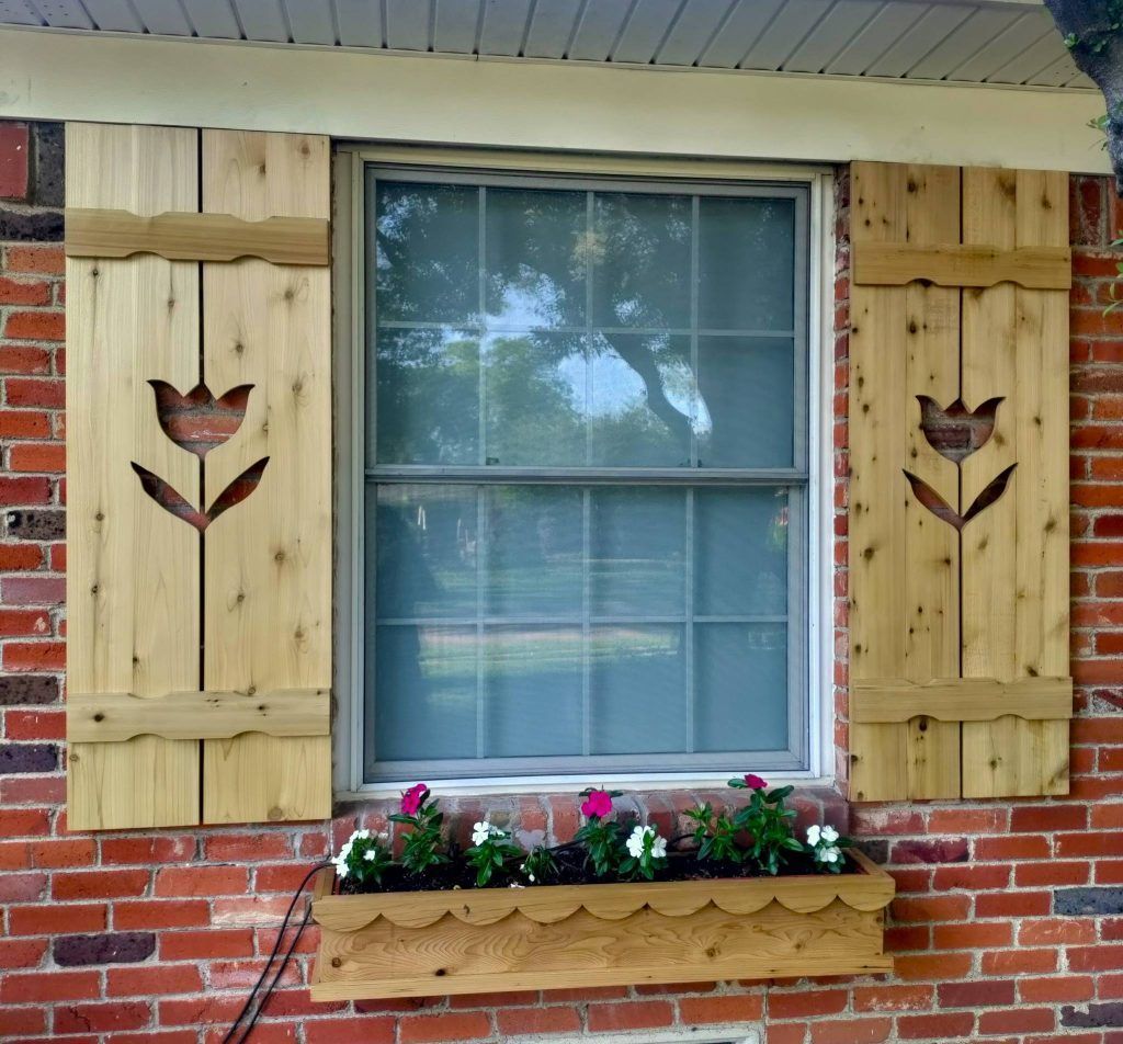 Wooden shutters with tulip cutouts frame a window over a brick wall; a scalloped-edge planter box below holds flowers.